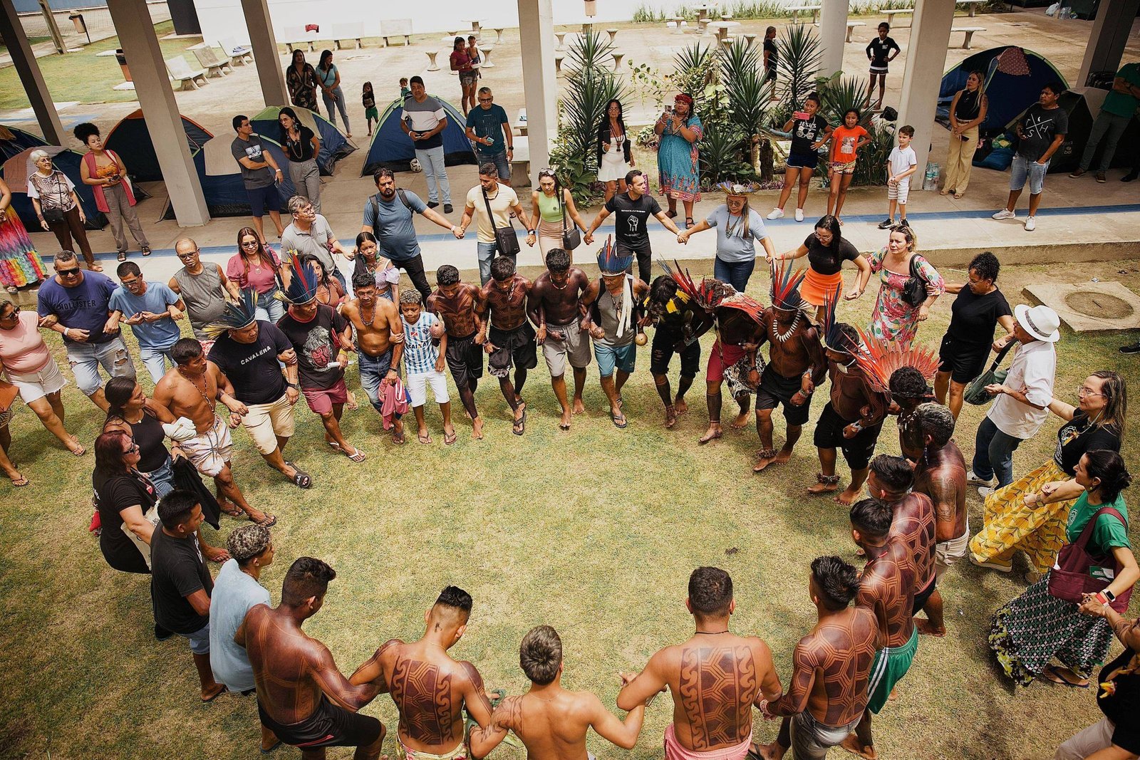 A imagem mostra um grupo de pessoas em um círculo, participando de uma dança ou celebração. No centro, há homens com pinturas corporais e penas na cabeça, enquanto outros ao redor seguram as mãos. O ambiente é ao ar livre, com tendas ao fundo e pessoas observando. A maioria dos participantes está vestida de forma casual, e há uma diversidade de idades e etnias entre eles.