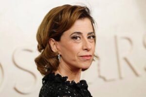 A imagem mostra uma mulher com cabelo castanho claro, preso em um coque, usando um vestido preto com detalhes. Ela está olhando para o lado, com uma expressão séria. O fundo é desfocado, mas parece ter a palavra