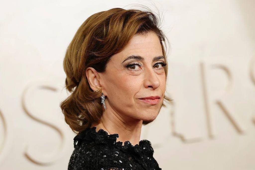 A imagem mostra uma mulher com cabelo castanho claro, preso em um coque, usando um vestido preto com detalhes. Ela está olhando para o lado, com uma expressão séria. O fundo é desfocado, mas parece ter a palavra