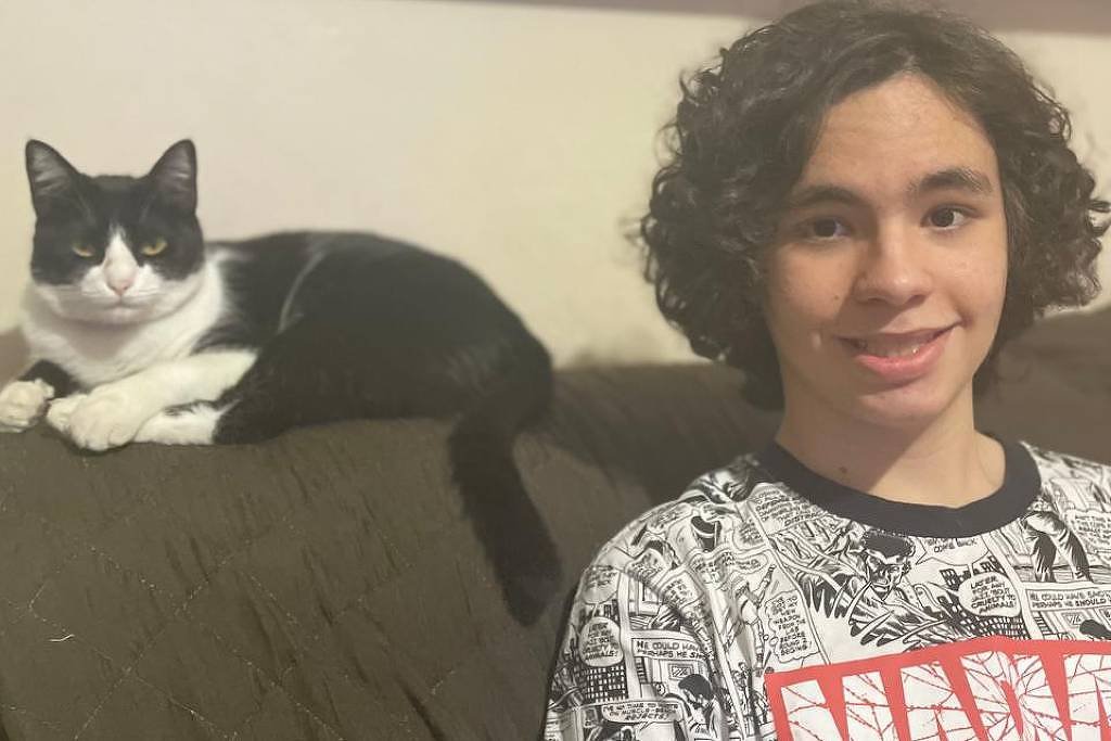 A imagem mostra um jovem sentado em um sofá, ao lado de um gato preto e branco que está deitado. O jovem tem cabelo cacheado e usa uma camiseta com estampas em preto e branco. O fundo apresenta uma prateleira com várias fotos emolduradas.