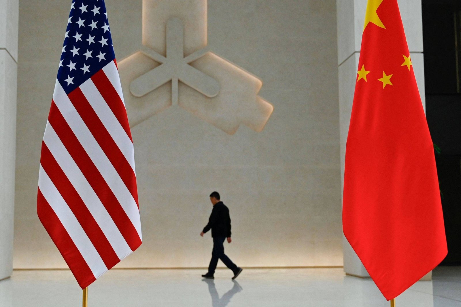 A imagem mostra as bandeiras dos Estados Unidos e da China em um ambiente interno. Um homem está caminhando ao fundo, entre as bandeiras, em direção a uma parede clara com um design abstrato. O ambiente é moderno e minimalista.