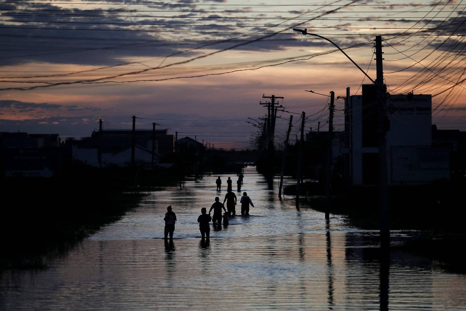 A imagem mostra uma cena de inundação ao entardecer, com várias pessoas caminhando em uma rua alagada. O céu apresenta nuvens e um tom de laranja e azul, refletindo a luz do sol poente. Ao fundo, há postes de eletricidade e edifícios parcialmente visíveis, com a água refletindo a luz e as silhuetas das pessoas.