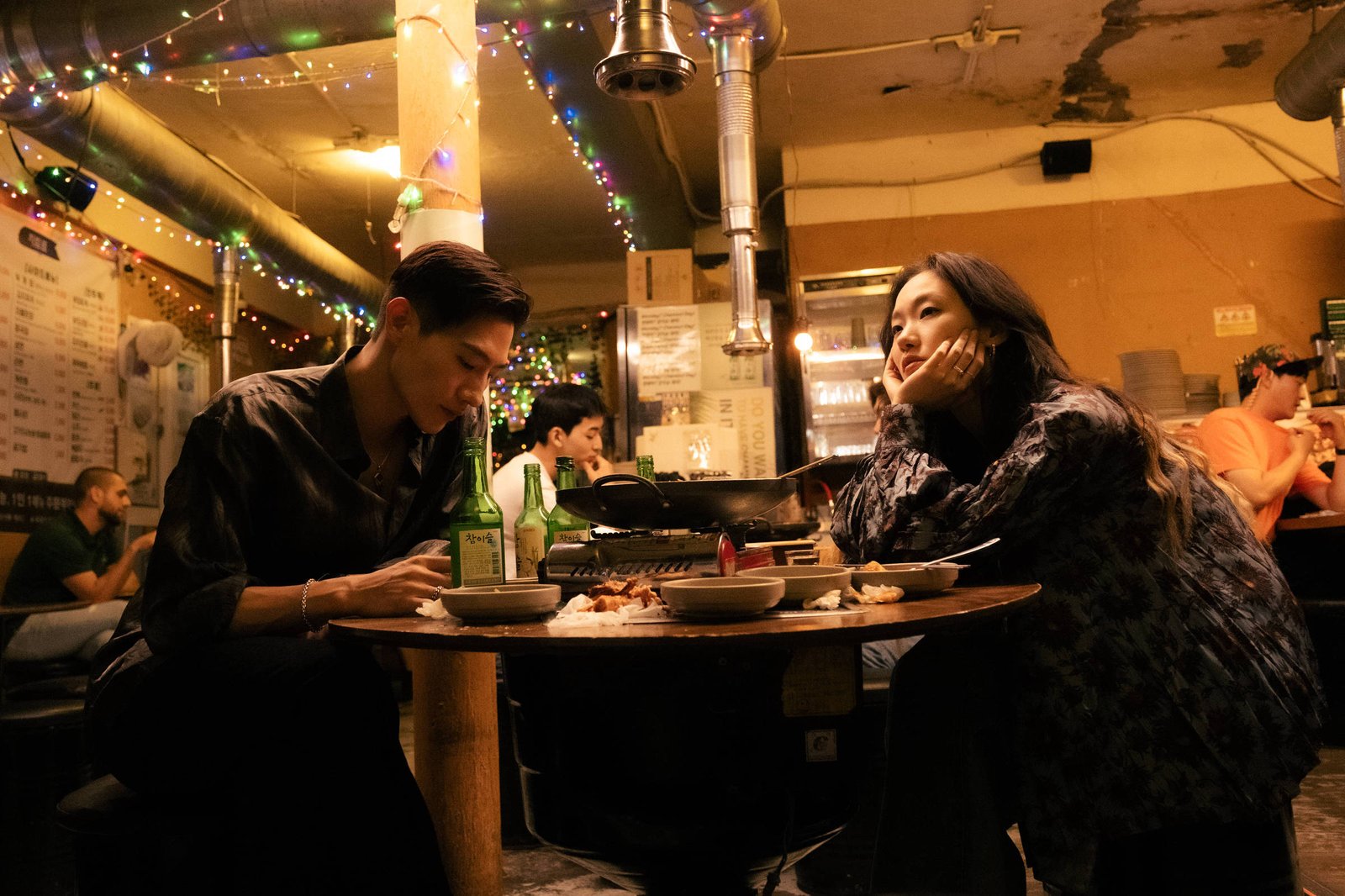 A imagem mostra um casal sentado em uma mesa em um restaurante. Eles estão em um ambiente com iluminação suave e decoração festiva, incluindo luzes coloridas. O homem, com cabelo curto e vestido de preto, está olhando para a mulher, que usa uma blusa escura e está apoiando o queixo na mão, sorrindo. Na mesa, há pratos de comida e uma panela quente. Ao fundo, outras pessoas estão sentadas e conversando.