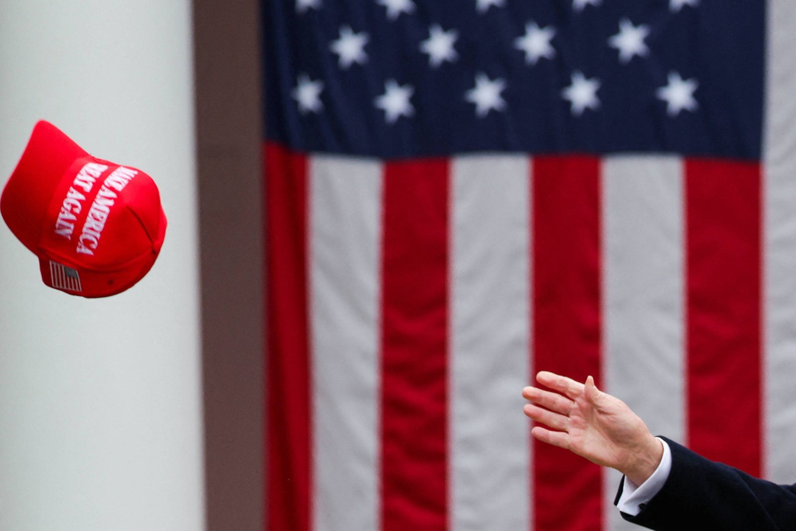 A imagem mostra um boné vermelho sendo lançado em direção à câmera, com uma mão visível no lado direito. Ao fundo, há uma bandeira dos Estados Unidos com listras vermelhas e brancas e estrelas brancas em um campo azul.