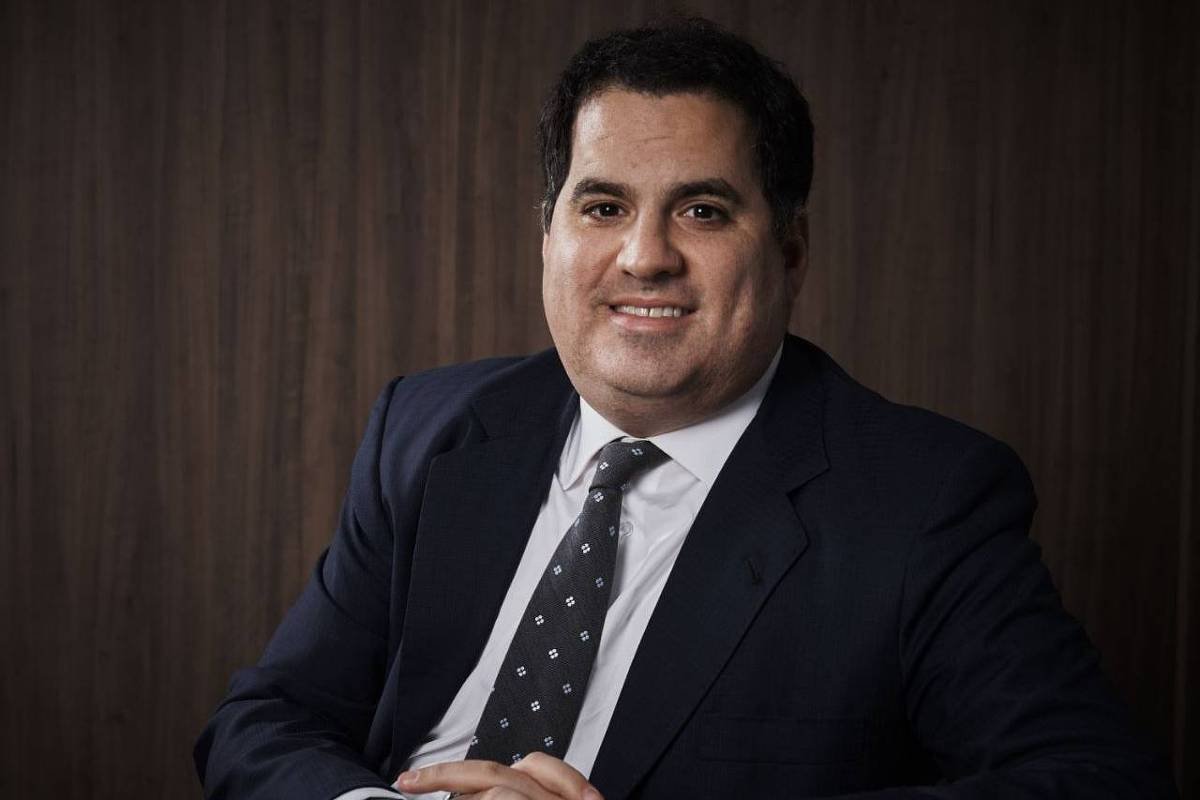 Luiz Gustavo Bichara é um homem branco com cabelo preto. Ele está usando um terno preto. Bichara sorri e cruza os dedos com as mãos apoiadas sobre uma mesa.