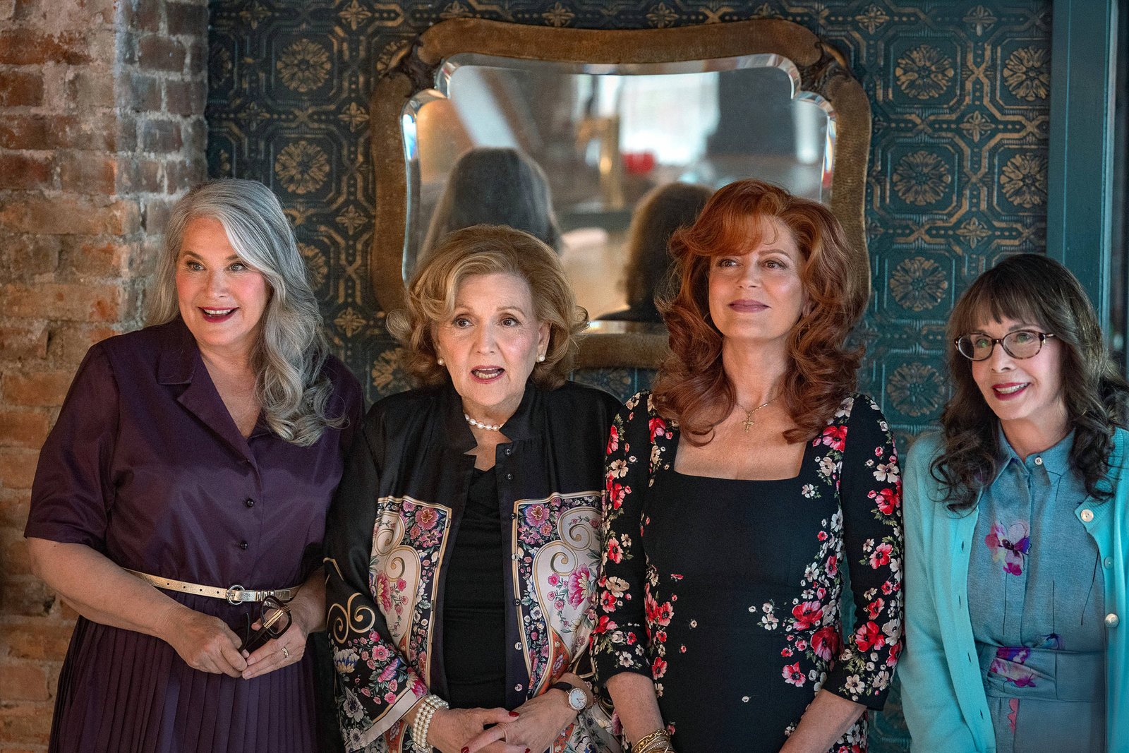 Quatro mulheres estão posando para a foto em um ambiente interno. A primeira mulher à esquerda tem cabelo longo e grisalho, vestindo um vestido roxo. A segunda mulher, ao lado dela, tem cabelo curto e castanho, usando uma blusa preta com um casaco decorado. A terceira mulher tem cabelo ruivo e usa um vestido preto com flores. A quarta mulher, à direita, tem cabelo castanho e usa uma blusa azul clara. Ao fundo, há uma parede de tijolos expostos e um espelho decorativo.