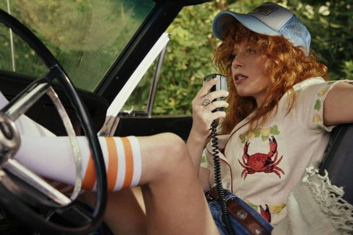 A imagem mostra uma mulher com cabelo cacheado e ruivo, usando uma camiseta clara com um desenho de caranguejo e um boné azul. Ela está sentada no banco de um carro antigo, com uma perna estendida e segurando um microfone. A cena é iluminada por luz natural, com vegetação visível ao fundo.