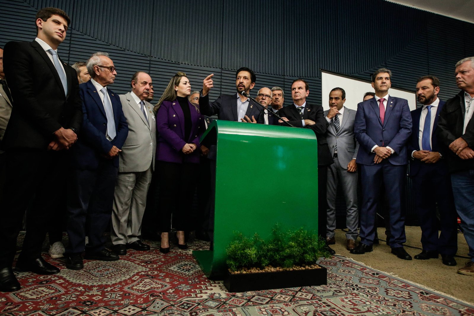Um grupo de políticos está reunido em uma coletiva de imprensa. No centro, um homem está falando ao microfone, enquanto os outros estão ao seu redor, ouvindo atentamente. O fundo é uma parede escura e há um painel verde à frente, com uma pequena planta na base. Os políticos estão vestidos formalmente, com ternos e gravatas, e a atmosfera parece ser de seriedade e atenção.