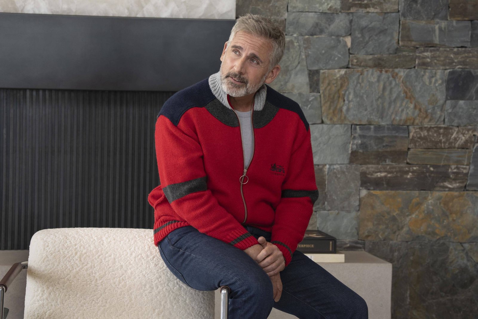 Um homem sentado em uma cadeira em um ambiente moderno. Ele está usando um suéter vermelho com detalhes em azul e uma camisa cinza por baixo. O homem tem cabelo grisalho e uma barba bem cuidada. Ao fundo, há uma parede de pedras e uma lareira. O ambiente é bem iluminado e minimalista, com alguns livros empilhados ao lado.