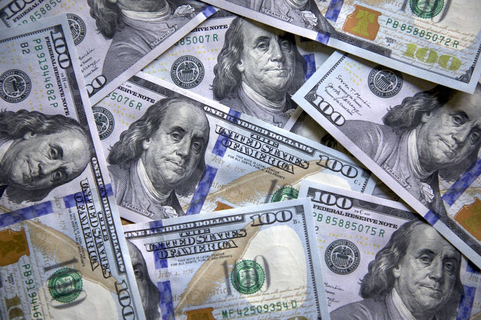 A imagem mostra várias notas de cem dólares dos Estados Unidos dispostas de forma sobreposta. As notas apresentam a figura de Benjamin Franklin e são predominantemente nas cores verde e azul, com detalhes em amarelo. O fundo é uma mistura das notas, criando um padrão visual.