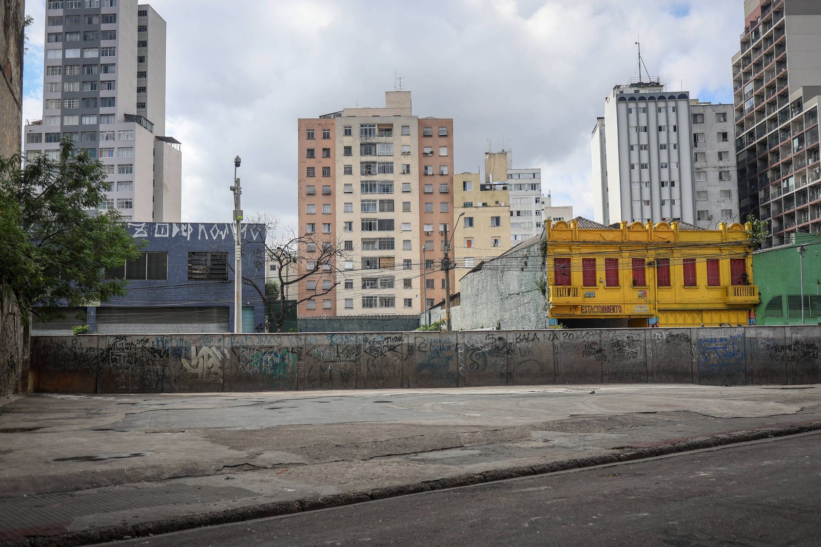 A imagem mostra uma área urbana com prédios altos ao fundo. À esquerda, há um edifício com uma fachada escura e uma inscrição em grafite. No centro, um espaço vazio com uma superfície de concreto, cercado por paredes grafitadas. À direita, destaca-se um prédio amarelo com janelas vermelhas, contrastando com os outros edifícios ao redor. O céu está nublado, sugerindo um dia sem sol.