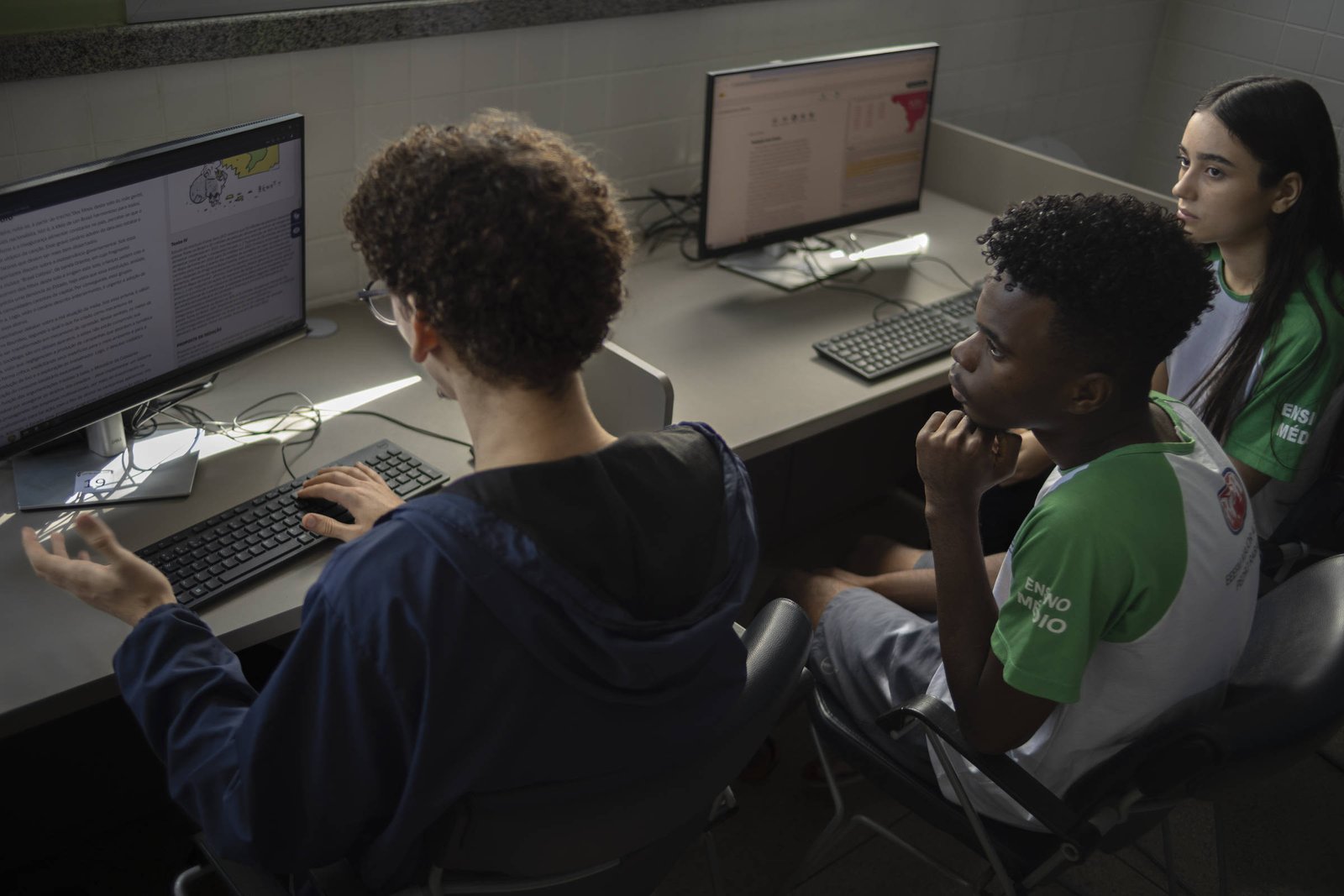 A imagem mostra três estudantes em uma sala de informática. Dois deles estão sentados em frente a computadores, enquanto um terceiro está de costas, gesticulando com a mão. Todos os estudantes estão usando camisetas com cores predominantes em verde e branco. As telas dos computadores exibem textos e gráficos. O ambiente é bem iluminado, com janelas ao fundo.