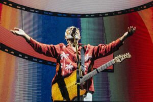 Um artista está se apresentando em um palco iluminado, com um fundo de telas que exibem padrões coloridos em espiral. Ele está usando uma jaqueta vermelha e segurando uma guitarra, com os braços abertos em uma pose expressiva. As luzes do palco criam um ambiente vibrante e dinâmico.