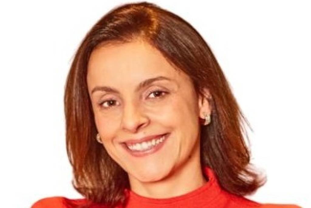 A imagem mostra uma mulher sorridente com cabelo castanho claro e liso, usando uma blusa vermelha de gola alta. O fundo é branco, destacando a figura da mulher que está com os braços cruzados.