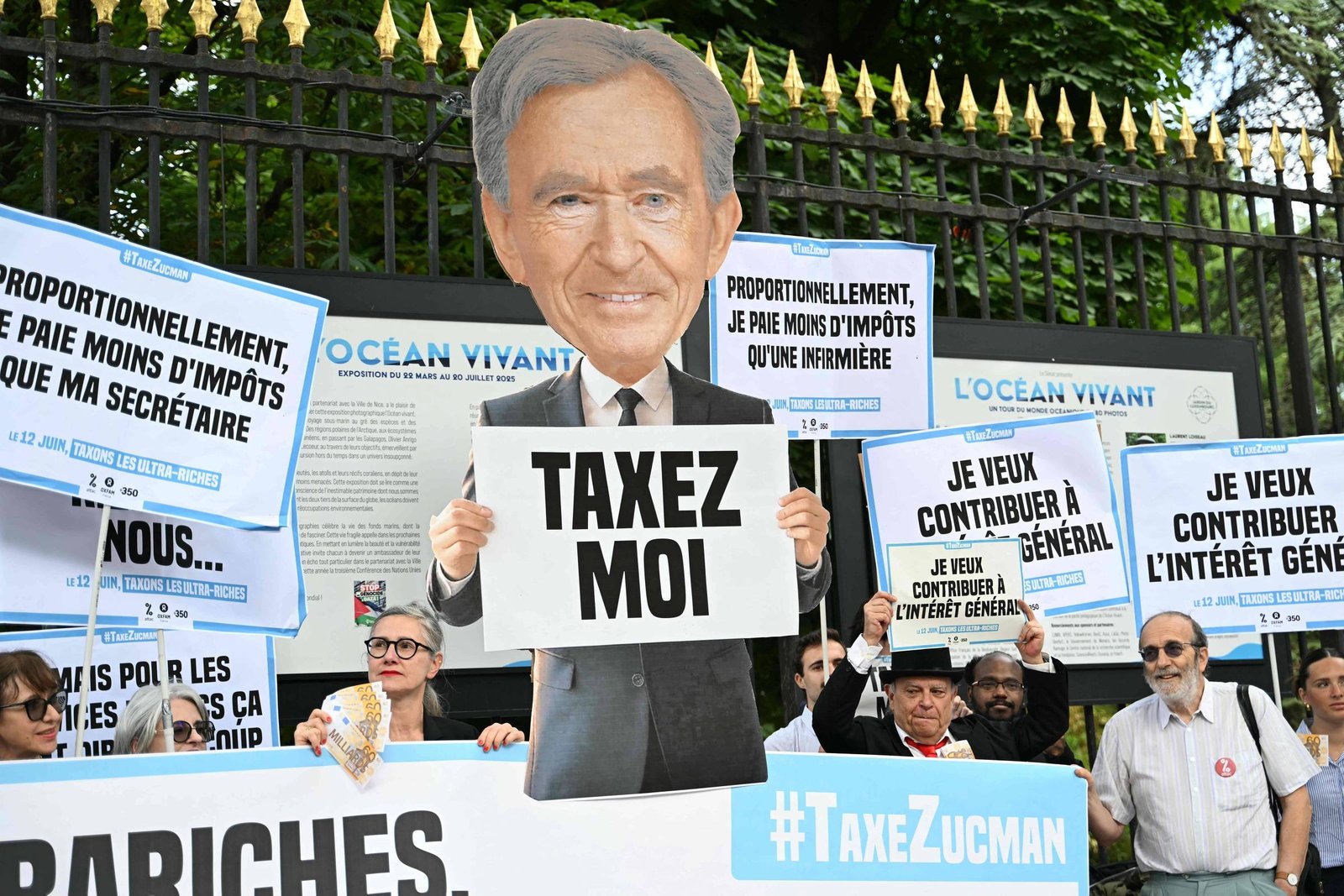 A imagem mostra um grupo de pessoas participando de um protesto. No centro, há uma figura em tamanho grande com a cabeça de um homem idoso, segurando um cartaz que diz