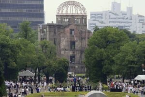 A imagem mostra o Memorial da Paz de Hiroshima, com a estrutura em ruínas ao fundo. Há uma grande quantidade de pessoas reunidas no parque, algumas em direção a um monumento em forma de arco sobre um lago. O ambiente é verde, com árvores ao redor e um céu nublado. Algumas pessoas estão usando capas de chuva, indicando que pode estar chovendo. O local parece ser um espaço de lembrança e homenagem.