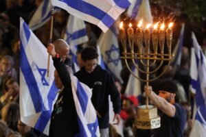 A imagem mostra um grupo de pessoas participando de uma celebração, segurando bandeiras de Israel. Um homem à esquerda levanta uma bandeira, enquanto outro à direita segura uma menorá acesa, que é um candelabro tradicional judaico com várias velas. Ao fundo, outras pessoas também estão visíveis, algumas segurando bandeiras. O ambiente parece festivo e iluminado.