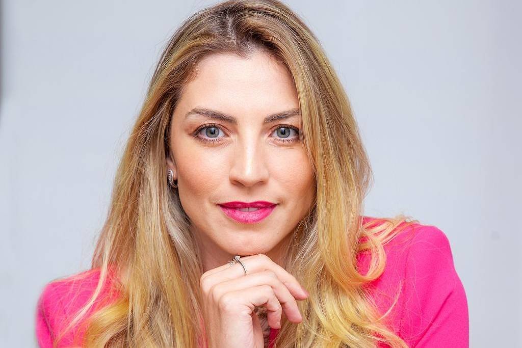 A imagem mostra uma mulher com cabelo longo e liso, usando um blazer rosa. Ela está sentada, com a mão direita apoiada no queixo, olhando diretamente para a câmera. O fundo é neutro, destacando a figura da mulher.