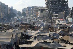 A imagem mostra uma vasta área coberta por tendas, indicando um acampamento. Ao fundo, há edifícios danificados, com um prédio em ruínas visível. O céu está claro, e a cena parece ser em uma cidade, com estruturas urbanas ao redor das tendas.