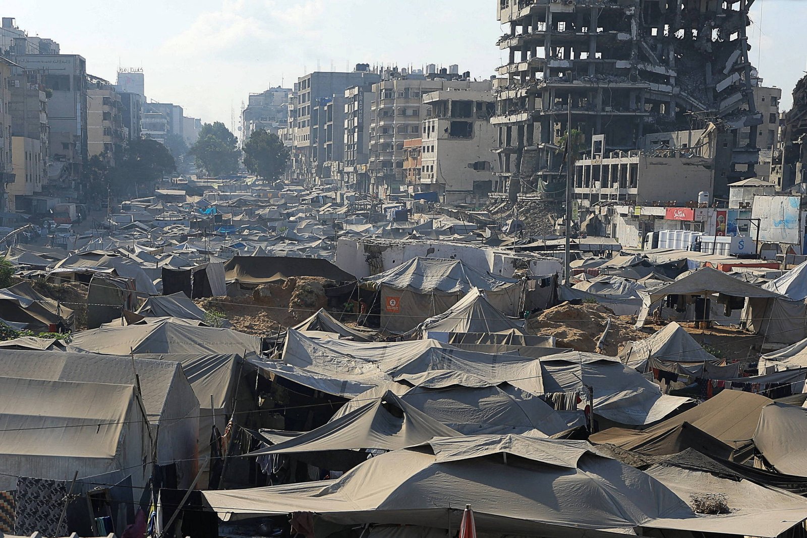 A imagem mostra uma vasta área coberta por tendas, indicando um acampamento. Ao fundo, há edifícios danificados, com um prédio em ruínas visível. O céu está claro, e a cena parece ser em uma cidade, com estruturas urbanas ao redor das tendas.