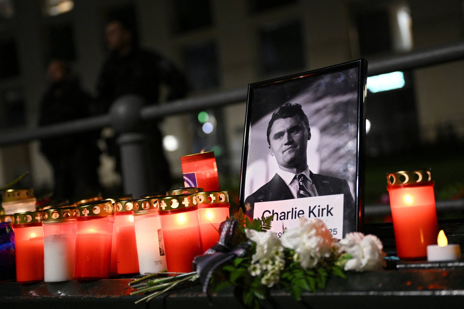 A imagem mostra um memorial com velas acesas e flores. No centro, há uma foto emoldurada de um homem, identificado como Charlie Kirk, com datas de nascimento e falecimento. As velas são predominantemente vermelhas e brancas, e o ambiente parece ser noturno, com algumas luzes ao fundo.