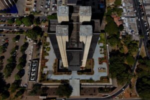 Vista aérea do prédio do Banco Central cercado por áreas verdes e ruas movimentadas. Ao redor, prédios compõem a paisagem urbana.