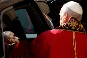 Homem com solidéu branco e capa vermelha com detalhes dourados está de costas, olhando para o reflexo de seu rosto em vidro escuro de um carro.