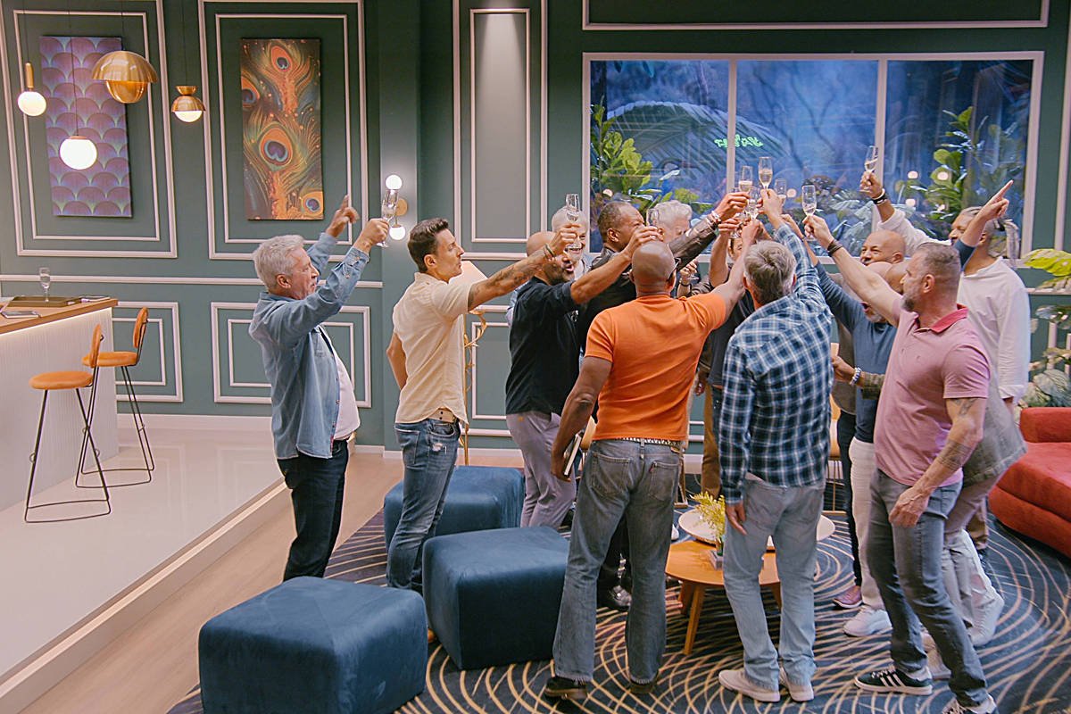 Um grupo de homens está reunido em um ambiente interno, levantando copos em um brinde. Eles estão em um espaço moderno, com paredes decoradas e uma grande janela ao fundo que mostra uma paisagem verde. O grupo é composto por diversas idades e estilos de roupas, e todos parecem estar se divertindo. O ambiente tem um sofá vermelho e algumas poltronas azuis.