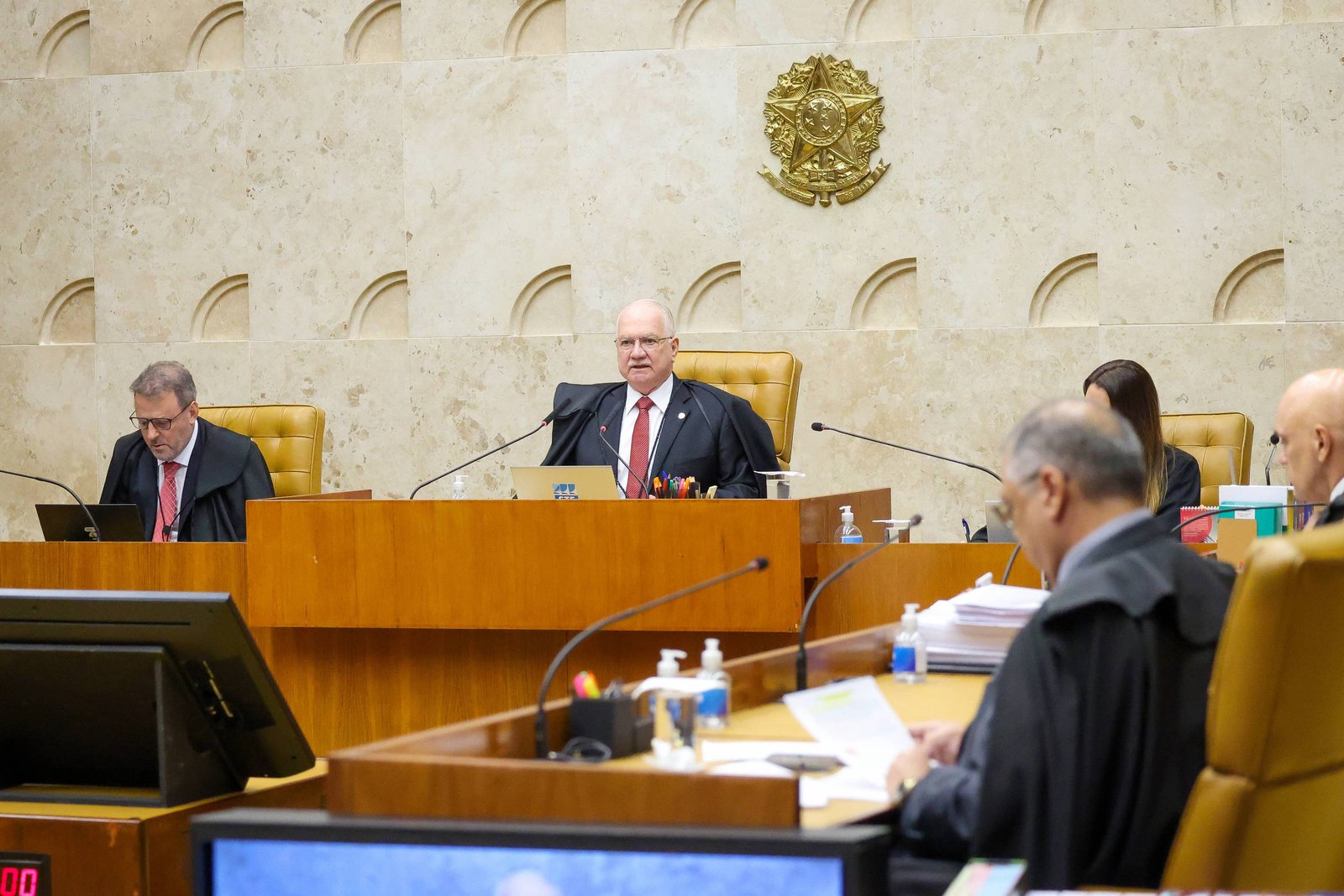 Quatro ministros do Supremo Tribunal Federal sentados em seus assentos no plenário. O ministro central fala ao microfone, com o brasão da República ao fundo na parede de mármore. Documentos, computadores e itens de higiene estão sobre as mesas.