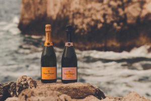 Duas garrafas de champanhe Veuve Clicquot, uma com rótulo amarelo e outra com rótulo rosa, estão posicionadas sobre pedras irregulares próximas ao mar. Ao fundo, ondas quebram contra uma formação rochosa grande e desfocada.