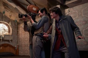 A imagem mostra três homens em um ambiente interno, possivelmente um quarto. Um dos homens está segurando uma câmera, enquanto os outros dois parecem estar em uma posição de alerta ou surpresa. O ambiente tem paredes com papel de parede vintage e há um ar condicionado visível. A iluminação é suave, e o cenário parece um pouco desordenado, com alguns tubos e equipamentos visíveis.