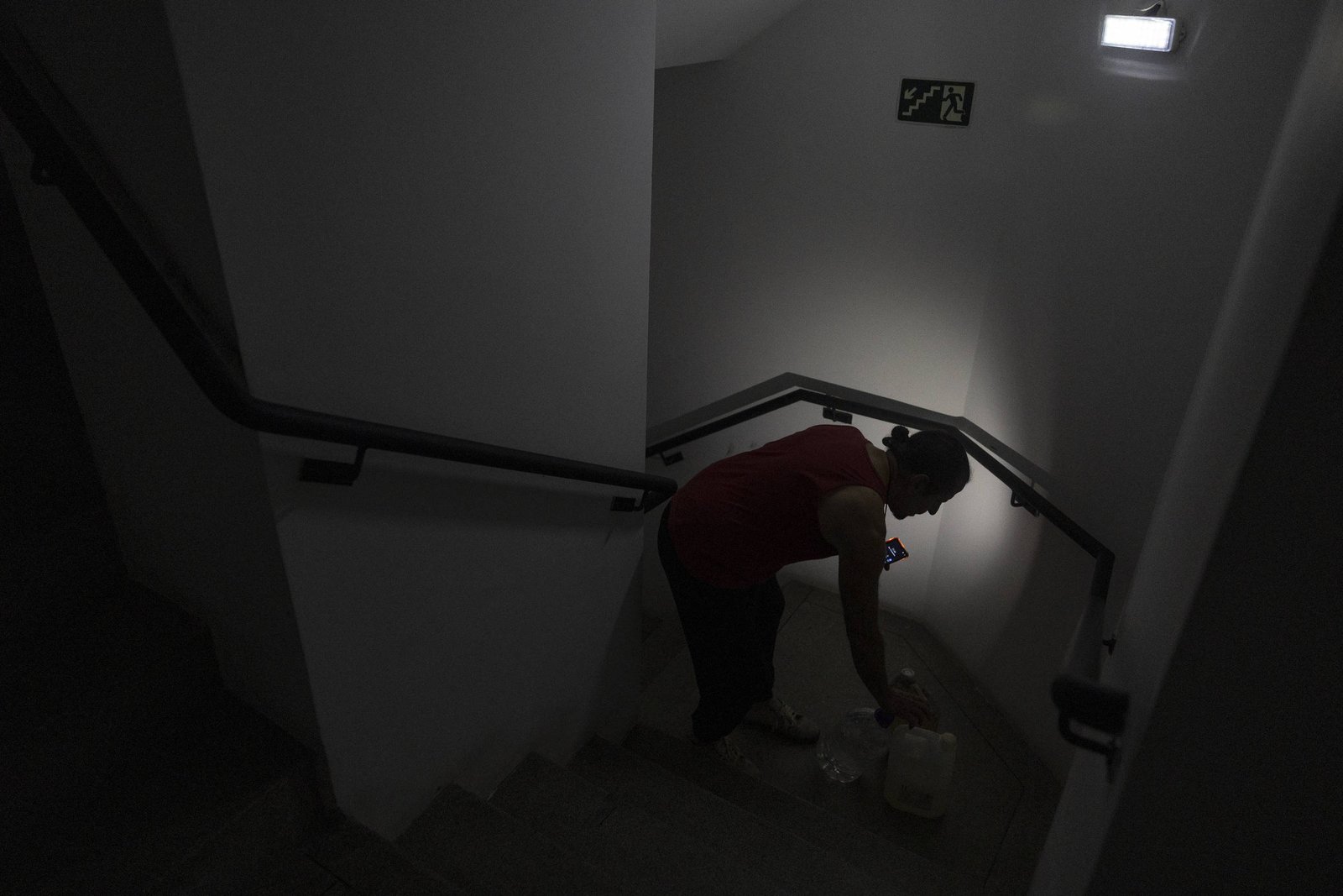 Mulher agachada recolhe água em balde no patamar de escada interna com iluminação fraca, usando lanterna de celular.