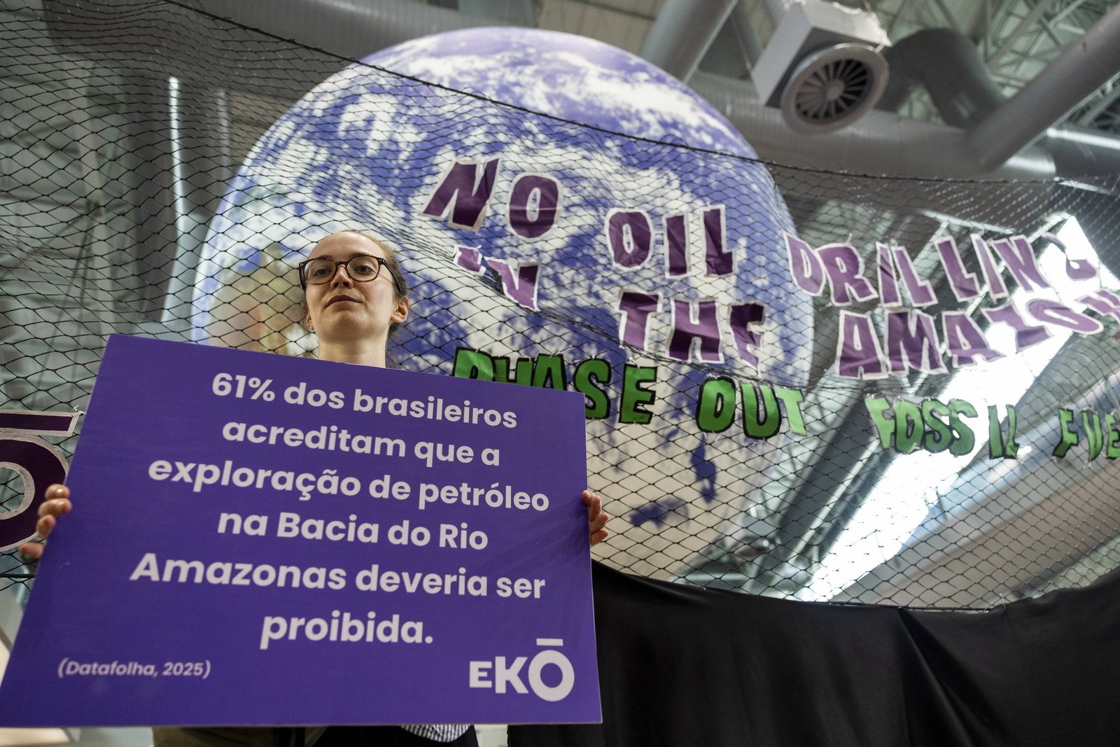 Pessoa segura cartaz com dados de pesquisa sobre proibição da exploração de petróleo na Bacia do Rio Amazonas, em frente a globo terrestre com mensagem contra perfuração de petróleo.