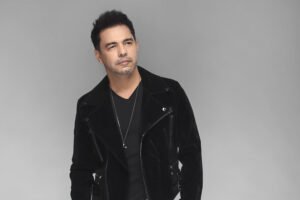 Homem em pé com mãos nos bolsos, veste jaqueta preta de veludo, camiseta preta, calça preta e botas pretas, com fundo cinza neutro.