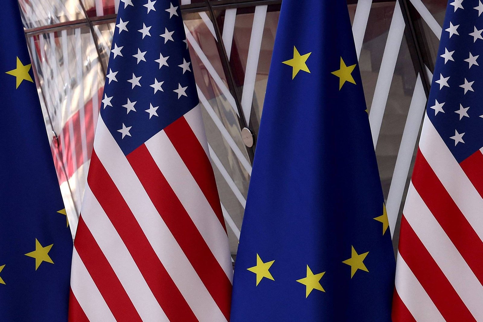 Quatro bandeiras alternadas dos Estados Unidos e da União Europeia alinhadas verticalmente. As bandeiras americanas exibem estrelas brancas em fundo azul e listras vermelhas e brancas, enquanto as bandeiras da União Europeia mostram estrelas amarelas em círculo sobre fundo azul.