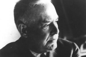 Homem idoso com cabelo curto e grisalho, vestindo terno escuro e camisa listrada, olhando para a direita em fundo neutro.