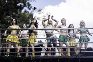 Sete jovens mulheres vestindo roupas esportivas coloridas, incluindo saias curtas amarelas e verdes, posam em um palco ao ar livre com céu azul e árvores ao fundo. Elas estão alinhadas atrás de uma grade metálica, algumas com as mãos na cintura e uma no centro com os braços erguidos segurando um microfone.