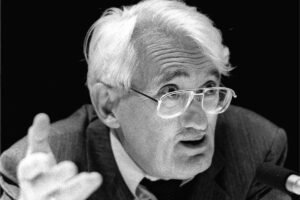 Homem idoso com cabelo branco e óculos transparentes fala gesticulando com a mão direita em frente a um microfone. Ele veste terno escuro, camisa clara e gravata preta, com fundo preto.