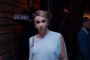 Pessoa com cabelo loiro preso, usando vestido branco sem mangas, posando em ambiente interno com iluminação baixa. Ao fundo, duas pessoas, uma carregando bandeja com copos e outra de terno azul.