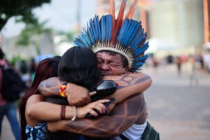 Homem indígena com cocar azul e vermelho abraça apertado duas mulheres em rua movimentada. Ele tem pintura corporal nos braços e expressão de emoção.