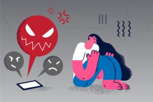 A imagem apresenta uma ilustração estilizada de uma pessoa sentada no chão, com as pernas abraçadas e a cabeça baixa, demonstrando tristeza ou angústia. Ao lado, há um balão de fala vermelho com um rosto zangado e expressões de raiva, além de dois rostos em tons de cinza, um com uma expressão neutra e outro com uma expressão de descontentamento. A paleta de cores é predominantemente em tons de azul, rosa e vermelho, com um fundo cinza.