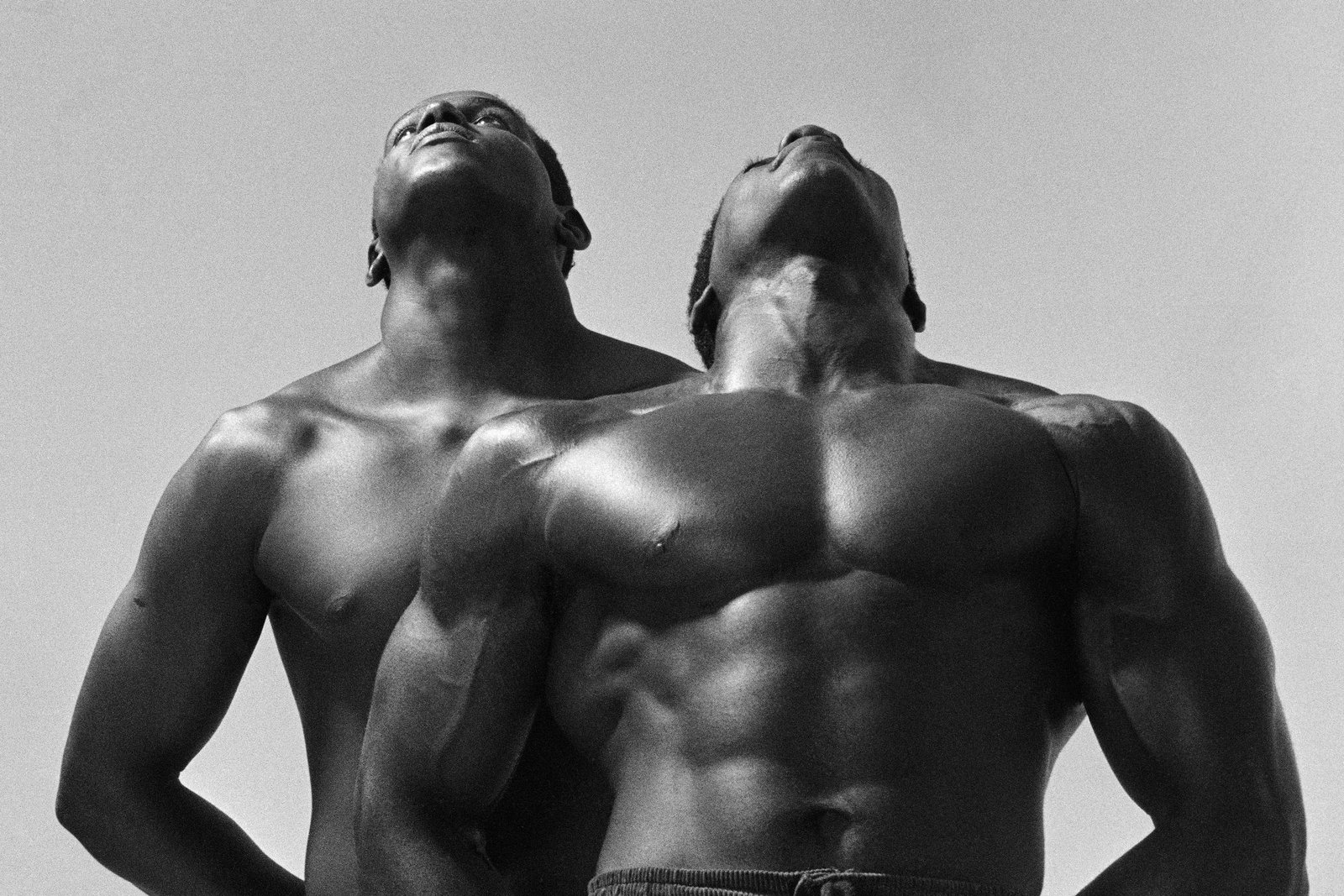Dois homens negros sem camisa exibem musculatura definida enquanto olham para cima contra um fundo claro e uniforme.