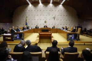 Ministros sentados em semicírculo no plenário do Supremo Tribunal Federal, com público em bancos à frente. Parede de fundo tem crucifixo e brasão da República, e bandeira do Brasil está à esquerda.