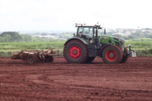 Trator verde com rodas vermelhas arando solo vermelho em campo aberto. Vegetação e área urbana ao fundo sob céu nublado.