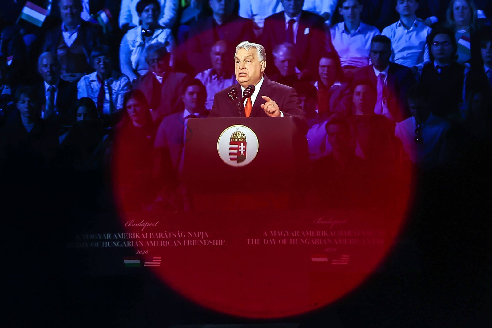Homem de cabelos grisalhos fala em púlpito com brasão da Hungria, cercado por público sentado em arquibancada ao fundo.