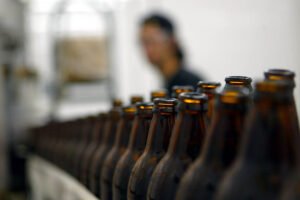 A imagem mostra uma linha de garrafas de cerveja marrons em primeiro plano, com um fundo desfocado onde uma pessoa está parcialmente visível. As garrafas estão alinhadas em uma superfície, e a iluminação é suave, destacando a transparência do vidro.