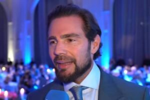 Homem de barba e cabelo escuro veste terno azul e gravata clara, falando ao microfone azul em ambiente interno com iluminação azul e público ao fundo.